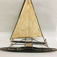 Front view of the model showing the triangular sail attached to wooden mast on dark wooden narrow canoe hull with outrigger.