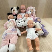 Front view of 9 dolls arranged on floor showing variety in types and outfits including realistic vinyl baby dolls and plush dolls.