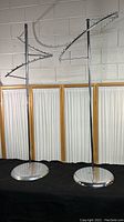 Two circular metal chrome clothing racks displayed side by side against a light-colored wall with curtains behind, showing full height and curved hook arms.