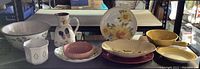 Wide shot of assorted ceramic dishware including bowls, platters, and pitcher on table surface showing variety in shape, size, and colors with floral motifs and painted decoration.
