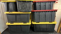 Five large plastic storage containers stacked and side-by-side, showing yellow and red lids and black bases. Containers appear used with weathering and dirt.
