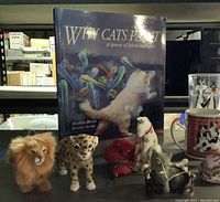 Single photo showing various small cat figurines including a lion, cheetah, and white cat figurine, with a book titled 'Why Cats Paint' behind them.