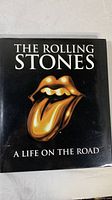 Cover of The Rolling Stones: A Life on the Road hardcover book with iconic tongue and lips logo in gold on black background.