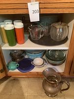 Wide view inside cabinet showing assortment of vintage kitchenware including colorful tall drinking cups, Pyrex measuring cups, glass ice bucket with silverplate lid, Corningware and other ceramic dishes, small teal teapot, and silverplate pitcher with ladle.
