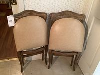 Front view of two wooden folding chairs with beige cushioned seats and cane backs against a wallpapered wall.