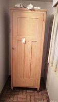 Front view of the vintage wooden armoire with a single closed door, natural wood finish, and a tag attached.