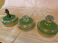 Set of three green glass bathroom containers with gold trim on a fabric surface. One container is a pump dispenser, the others are covered jars. All are shown closed.