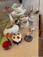 Photo of assorted white porcelain and ceramic vases, urn, decorative boxes, a metal candle holder, and red floral painted small pitcher arranged on table.