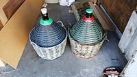 Two large glass demijohn bottles with woven wicker coverings, placed on concrete floor in garage area.
