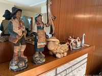 Photo showing large ceramic figurines of a man and woman in traditional attire with additional small animal figurines on a wooden mantelpiece