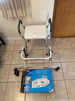 Shower chair with white perforated seat and padded armrests, two black canes, and boxed Carex hinged toilet seat riser on tiled floor.