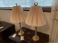 Pair of two side table lamps with cream pleated lampshades, brass bases and clear decorative glass column detail. Visible cords and tags on lamps.