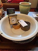 Photo showing white ceramic basin holding two leather straps, a small vintage clock in a brass frame case, and black slide viewer. Green ceramic pot is behind basin.
