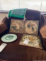 Four decorative ceramic plates with original art designs arranged on a brown couch showing various mythological and classical themes.