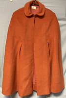 Front view of an orange wool cape or swing coat with rounded collar and slit armholes.