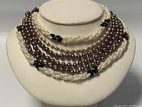 Three faux pearl necklaces displayed on a white bust: multi-strand white and black choker, long dark purple lariat, and twisted white with black beads.
