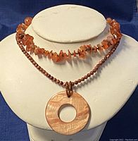 Photo showing two necklaces on display: one with orange amber chip beads and small round beads, and one with a large circular flat pendant and small round reddish-brown beads.
