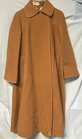 Front view of camel brown wool coat with collar, long silhouette, and pockets, hanging on hanger over white fabric background.