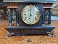 Front view showing the clock face, key on the floor, black wooden case, marble-look accents, fluted green columns, metal floral decorations, and brass cabriole feet.