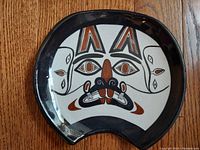 Front view of curved pottery with beaver mask design in black, white, and rust colors on wooden floor.