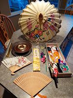 Full view of Eastern Asian collectibles group including parasol standing open in background, two folding fans, two decorative paddle fans in box, Japanese lidded bowl, and calendar scroll laid out on table.