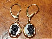 Front view of both cameo earrings on wooden surface showing oval black-on-white cameos with gold plated frames and leverback hooks.