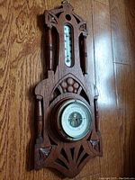 Front view of oakwood encased vintage German barometer and thermometer instrument showing detailed wood carving, thermometer at top, and barometer dial below.