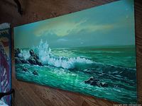 Full view of the rectangular oil painting depicting a seascape with waves crashing on rocks, placed on a wood floor.