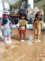 Three vintage First Nations female form traditional dress figurines standing on wooden bases on a wooden floor, detailed with leather, beadwork, fur, and embroidery.