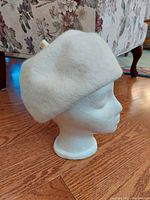 Side view of white felt beret showing its rounded shape and soft texture, placed on white mannequin head.