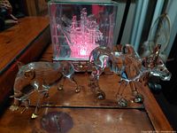 Three clear glass animal figurines (bull, unicorn, pig) on wooden surface with acrylic fibre optic crystal tall ship in back producing pink light