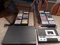 Panasonic cassette deck with cassette inserted, Daiwa DVD player, and two racks filled with vintage rock music cassette tapes on a table.