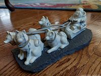 Side angled view of the full sculpture showing Inuit figure on sled and four dogs, with leather reins on granite mount.