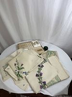 Photo showing several embroidered linens stacked on a small round white table, some partially still in packaging, with a green spherical size reference object