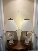 Front view of both rooster table lamps on wooden surface against wall.