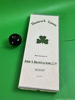 White box labeled Shamrock Linens by John S. Brown & Sons Ltd, Belfast, Ireland, next to a black round object used for scale. Shows packaging and branding.