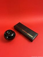 Black rectangular presentation box with gold Ferrari Formula logo shown closed next to glossy spherical reference object