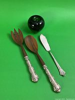 Three serving utensils including two wood-handled salad servers with sterling silver decorative handles, one silver hallmarked butter or fish serving cutting utensil, all on green background which also displays a green apple for size reference