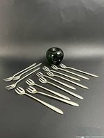 Twelve forks arranged on black surface with a green glass apple for size reference.