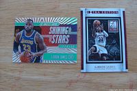 Two LeBron James basketball cards placed side by side on a wooden surface. Left card is Panini Illusions Shining Stars Emerald featuring LeBron James in Lakers uniform. Right card is Panini Extra Edition showing LeBron James in Cleveland Cavaliers uniform.