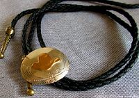 Close-up of the oval medallion centerpiece showing the brass cowboy hat and engraved nickel silver base along with the braided black cord and brass cord tip ball.