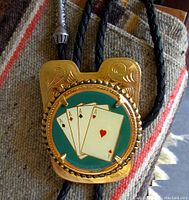 Front view of vintage bolo tie slide showing the 4 Aces design in gold-tone metal on a green background with black braided leather cord.