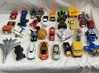 Entire assortment of 24 toy vehicles laid on a white fabric background showing multiple types and colors including cars, trucks, planes and motorcycles.