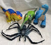 Four large toy figures including a blue and orange Tyrannosaurus rex, a green and beige Spinosaurus, a blue Brachiosaurus, and a black spider with long legs placed on a white surface
