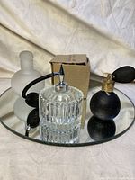 Photo of three vintage perfume bottles and atomizers on a round beveled glass mirror base, showing clear ridged atomizer, black atomizer with bulb pump, and frosted glass bottle