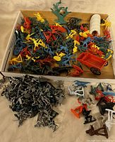 Box filled with assorted small vintage plastic toy soldiers, cowboys, and accessories in multiple colors including red, yellow, blue, green, orange, gray, black, and white.