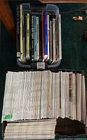 Top view of plastic tote filled with various books standing upright showing titles and multiple books and magazines laid flat beneath it, showing spine texts.
