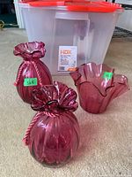 Three cranberry glass vases shown in front of the storage bin. Two are pouch-shaped with twisted glass rope details and one has a wide fluted edge.