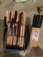 Wooden knife block with multiple knives inserted, placed in plastic bin next to second wooden knife block also with knives.