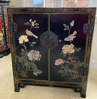 Front view of closed cabinet showing black lacquer finish, bird and floral appliqué motifs, metal hinges and clasp.
